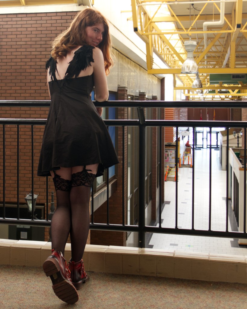 A person with long, wavy red hair leans against a balcony railing in a black dress with feathered details, showcasing fishnet stockings and lace garters, with a hallway blurred in the background.